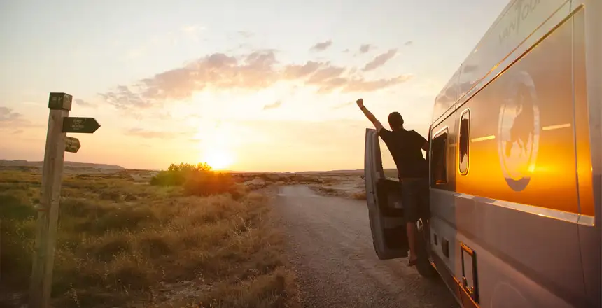 Fahrer in Tür eines Campervans streckt Arm Richtung Sonnenuntergang