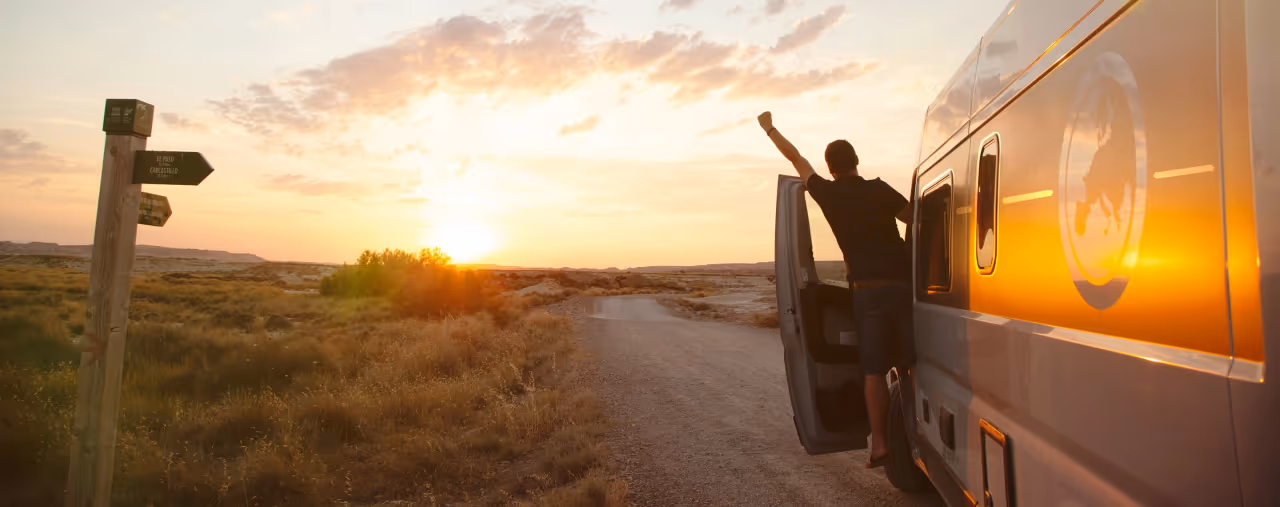Fahrer in Tür eines Campervans streckt Arm Richtung Sonnenuntergang