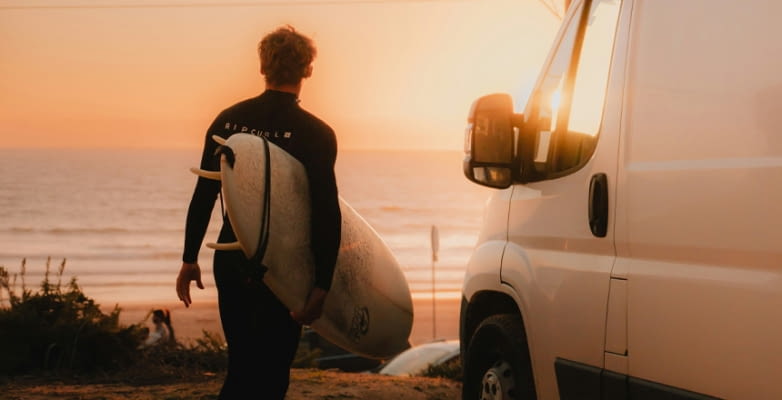 Surfer mit Brett im Sonnenuntergang neben Campervan mit Blick zum Meer